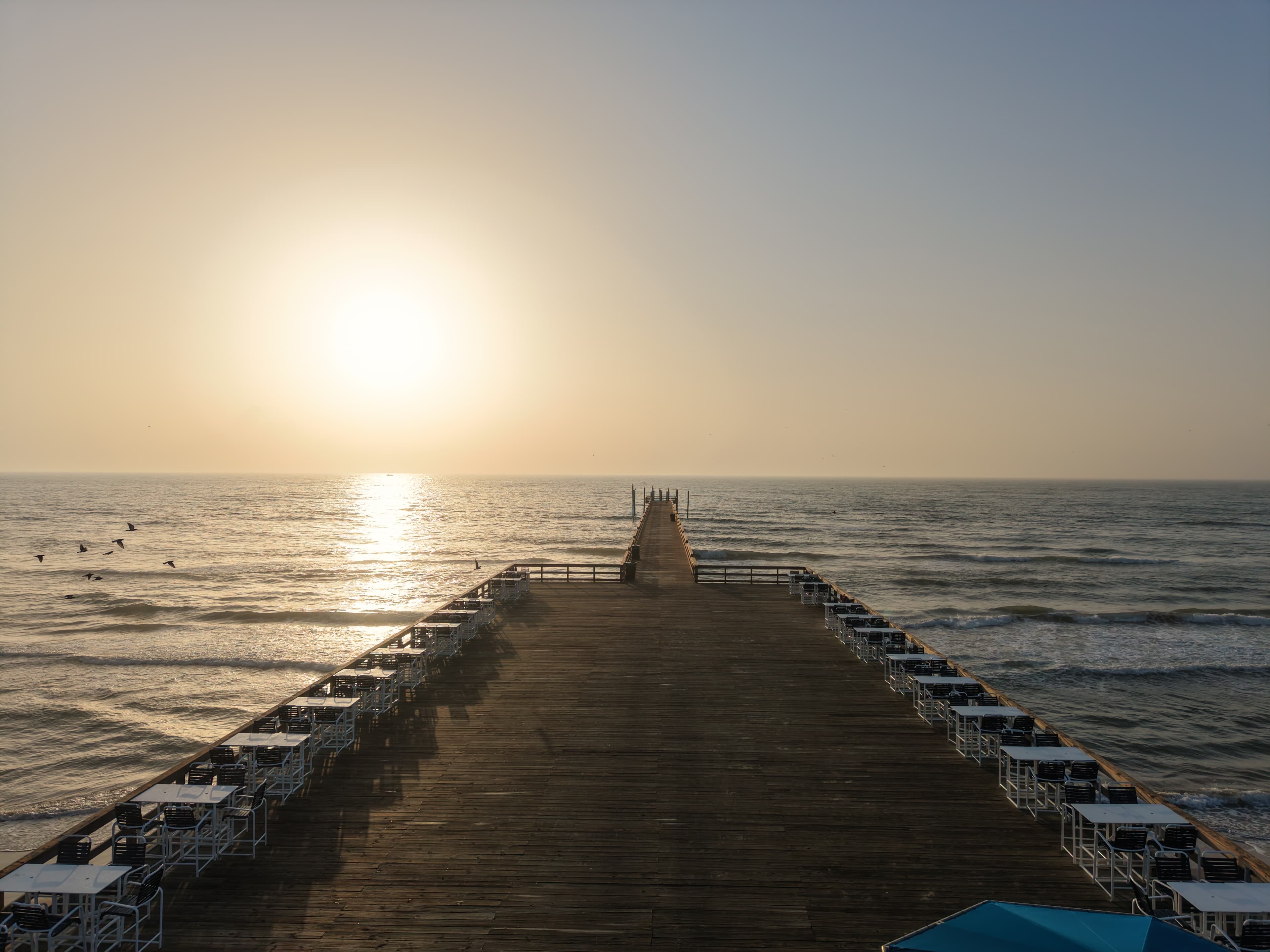 Wooden pier with dining tables stretching into the ocean under a bright hazy golden sunset.